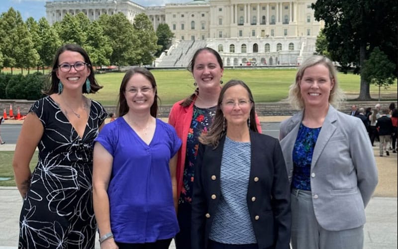Five women smiling and posing in front of the U.S. Capitol building on a sunny day, with trees and grass visible in the background.
