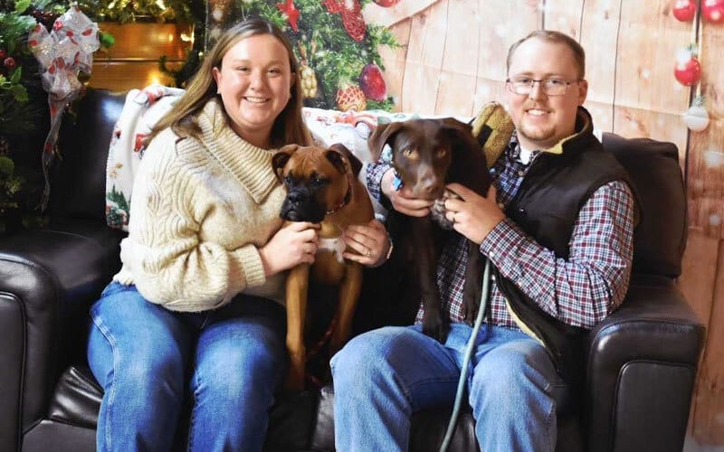 A smiling couple sits on a black couch with Christmas decorations behind them, each holding a dog—one brown and one dark brown. The woman wears a cream sweater and jeans; the man wears glasses, a plaid shirt, and a vest.