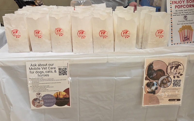 A table covered with a white cloth holds two rows of small paper popcorn bags. Signs on the front promote mobile vet care for pets and provide information about Red Oak Animal Hospital.