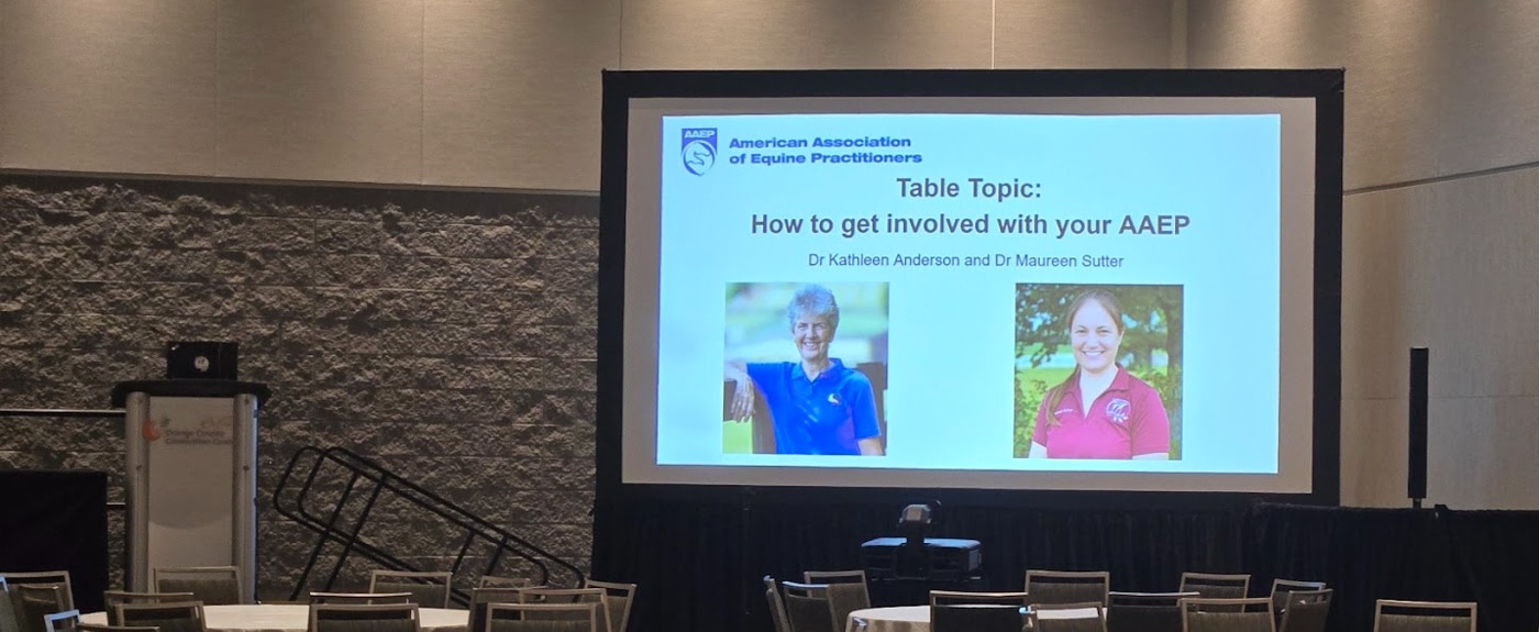 A conference room with a large screen displaying a presentation titled "How to get involved with your AAEP" by Dr. Kathleen Anderson and Dr. Maureen Suter, featuring their photos and the American Association of Equine Practitioners logo.