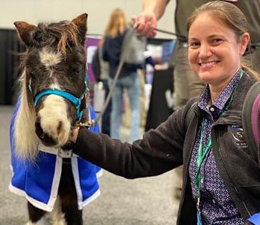A woman crouches and smiles next to a small horse wearing a blue coat and halter. The Veterinarian is gently petting the horse, with people visible in the blurred indoor background of an Animal Hospital.