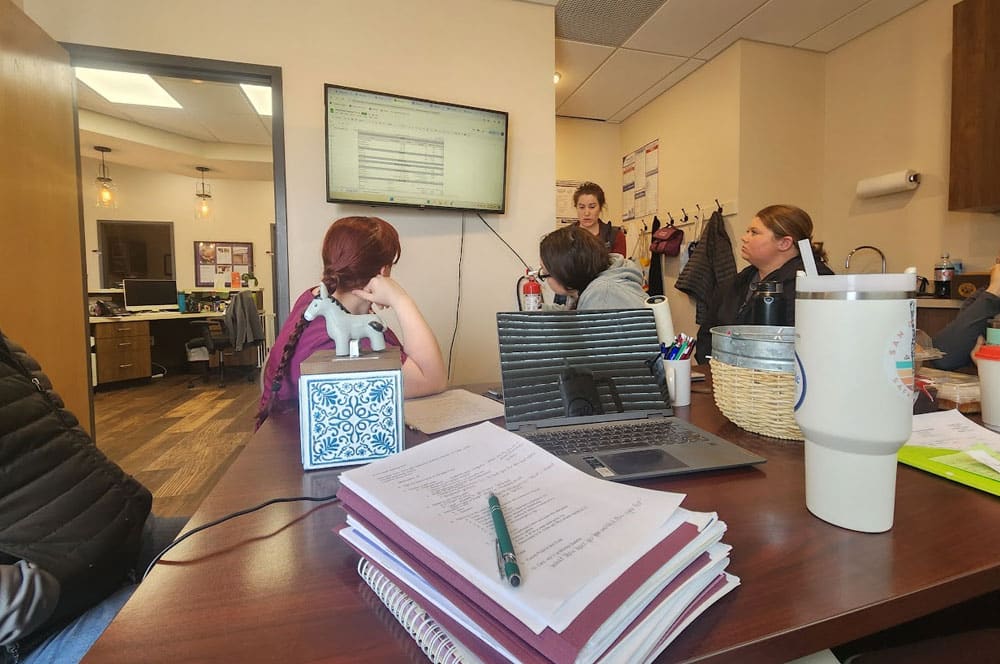 Several people sit around a meeting table with laptops, notebooks, and drinks. A woman stands near a screen displaying a spreadsheet, while others listen attentively. Office supplies and coats are visible in the background.