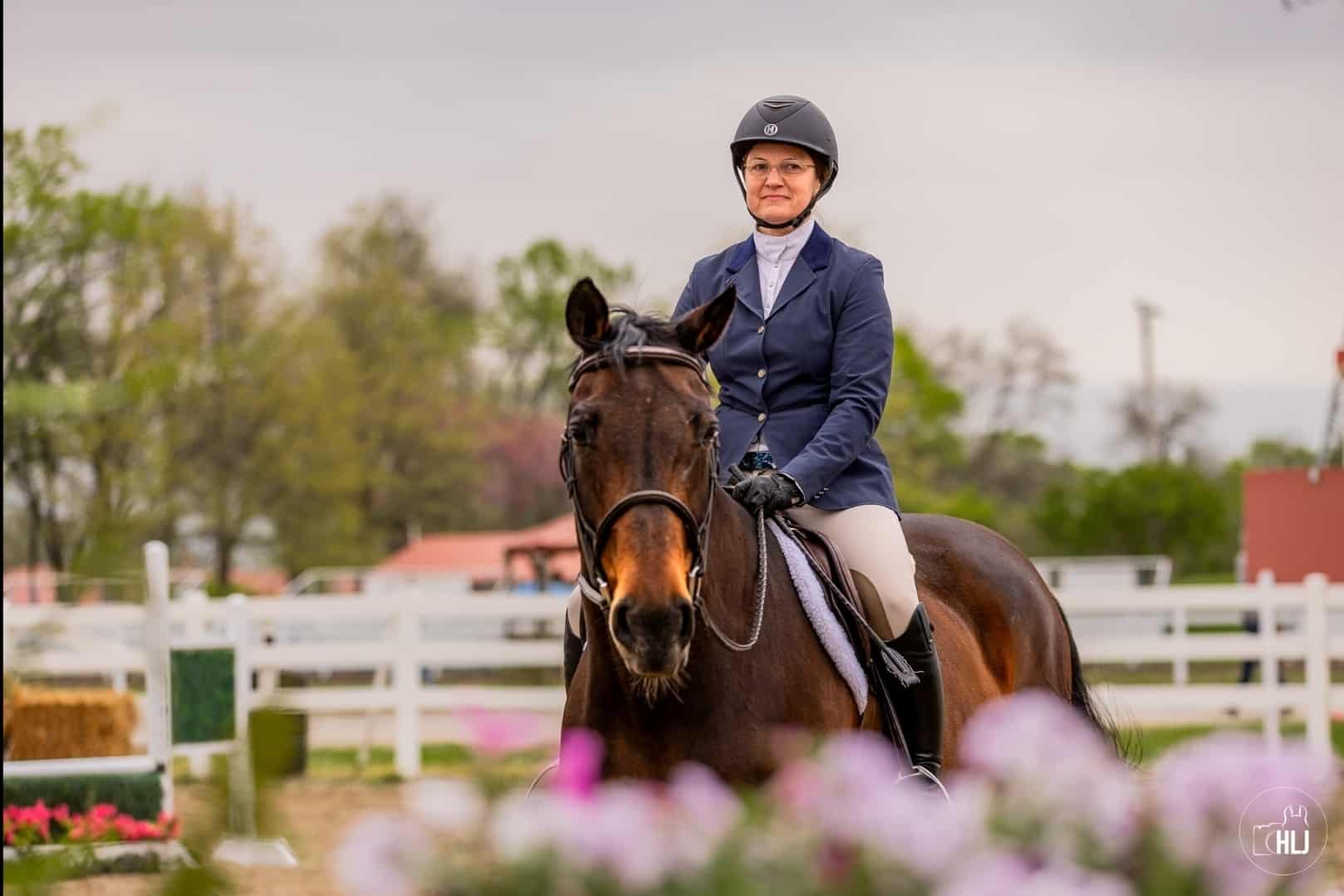 A rider in a navy jacket, white breeches, and a black helmet sits on a brown horse in an outdoor arena near a pet clinic, surrounded by white fences and blooming flowers, with trees and buildings in the background.